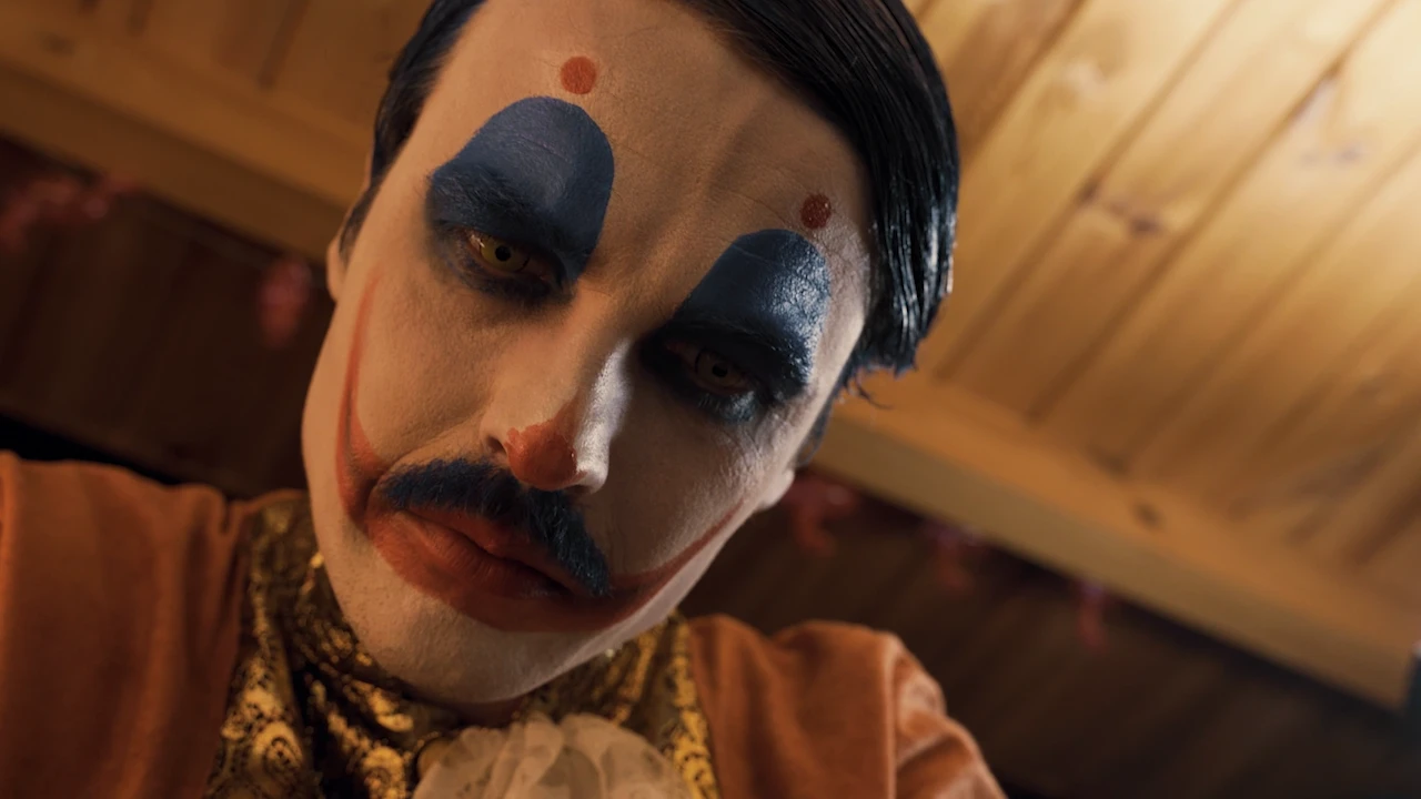Großaufnahme des Killerclowns Mitchell mit blauem Augen-Make-up und Schnurrbart, er blickt von oben herab.