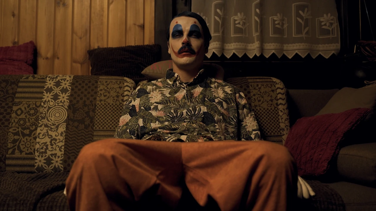 Der geschminkte Clown sitzt breitbeinig in Hawaiihemd und orangefarbener Hose auf einem alten Sofa.