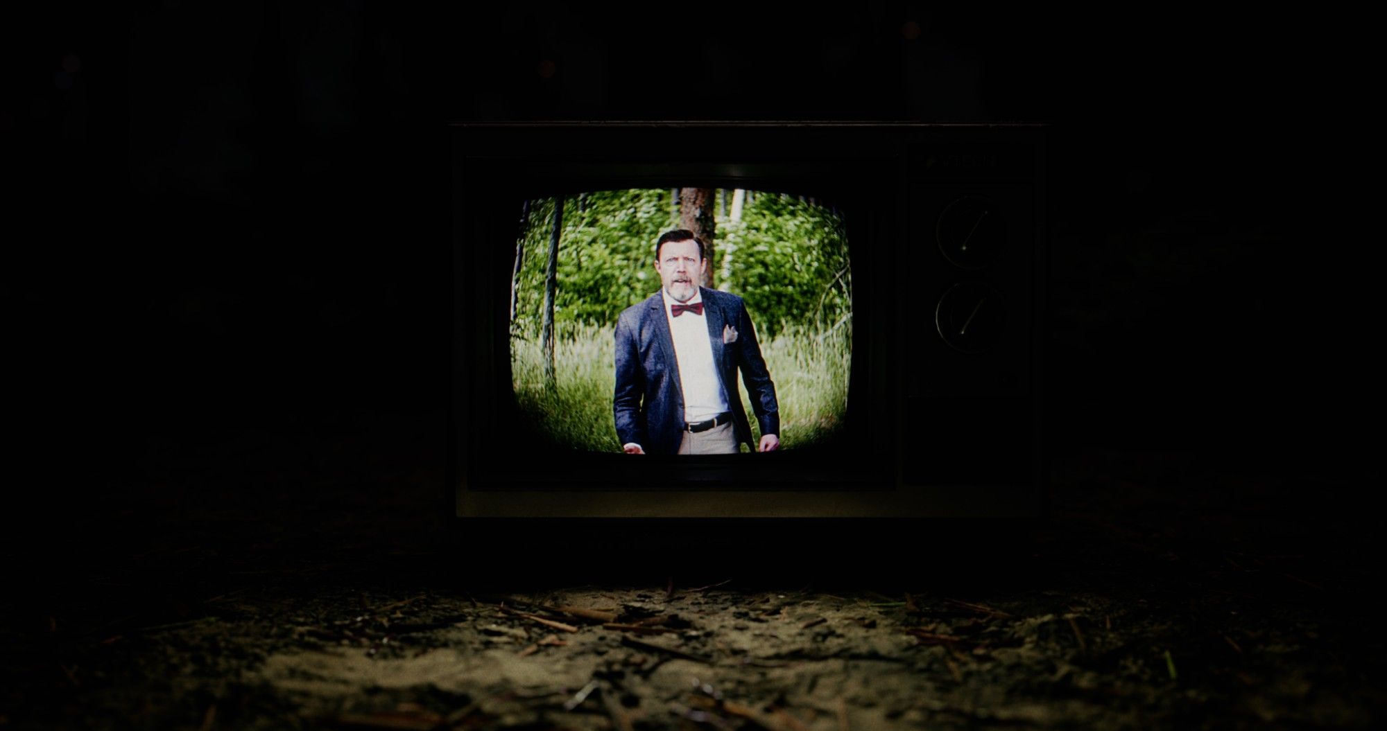 Ein alter Röhrenfernseher steht nachts im Wald, auf dem Bildschirm ein Mann mit Fliege und Sakko.