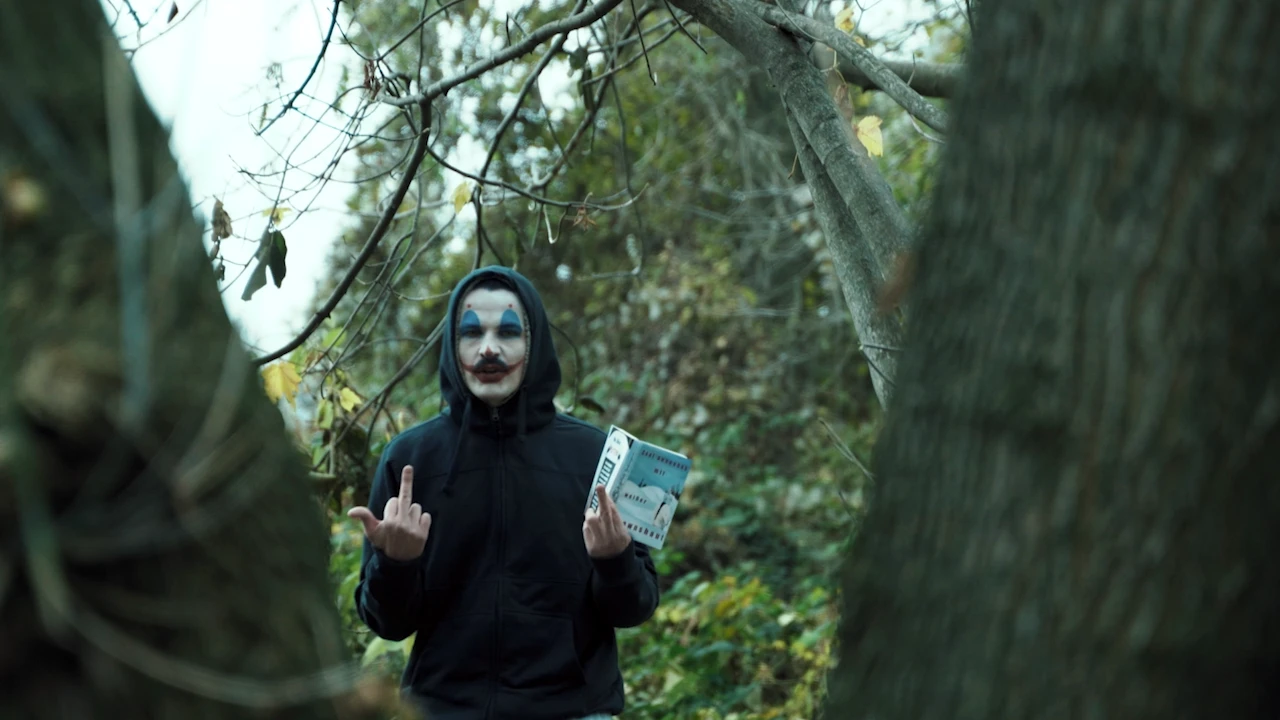 Ein Clown im Kapuzenpulli zeigt im Wald den Mittelfinger und hält eine Packung in der Hand.