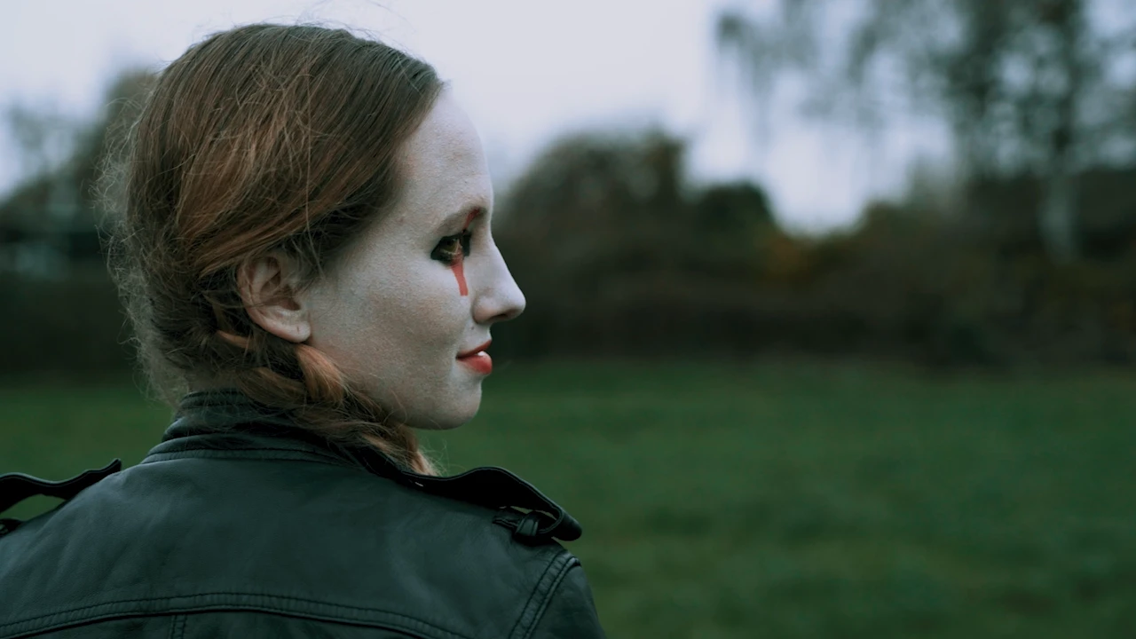 Eine Clownin mit weißer Schminke und roter Träne im Profil, im Hintergrund eine graue Wiese.