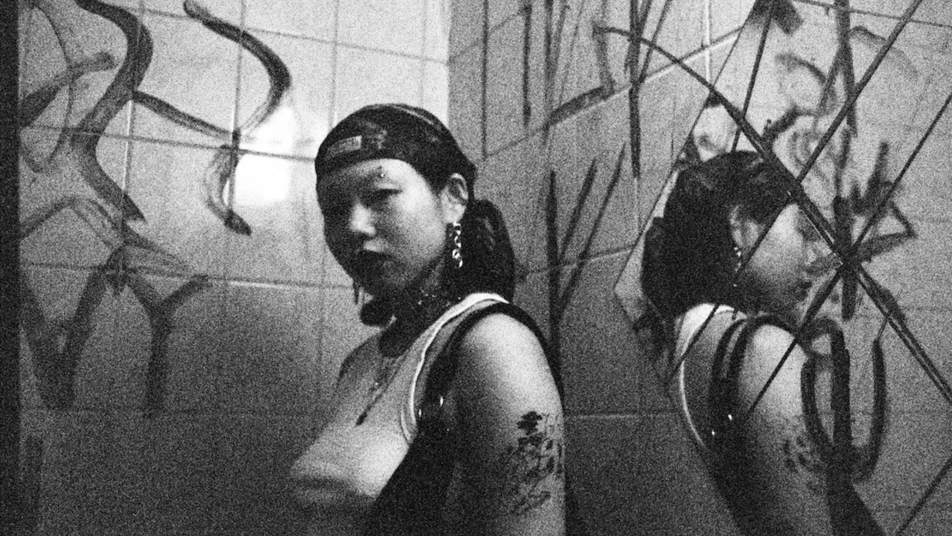 Schwarzweißfoto: Eine junge Frau mit Kopftuch und Piercings lehnt an einer bemalten Fliesenwand, daneben ihr Spiegelbild in einem gesprungenen Spiegel.